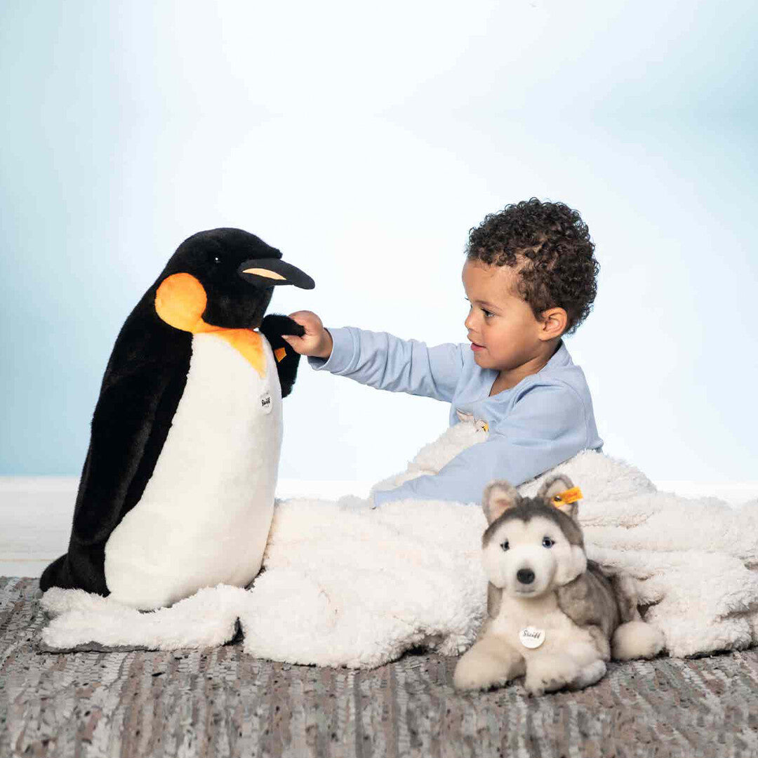 Een kind speelt met een grote pluchen pinguïn, met de Steiff pluchen knuffelhond husky Bernie ernaast.
