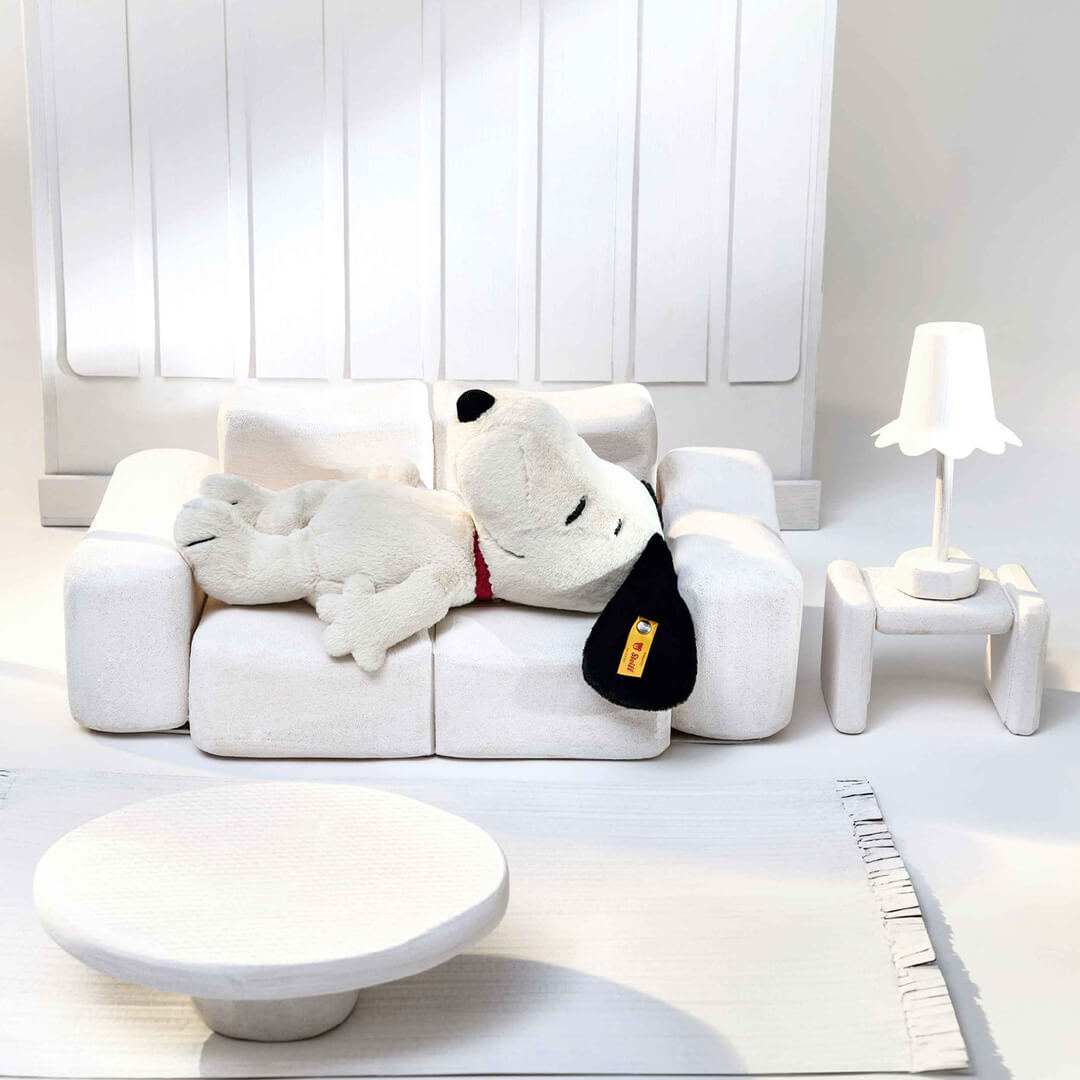 Steiff pluchen Snoopy knuffel liggend op een witte miniatuur bank in een witte kamer.
