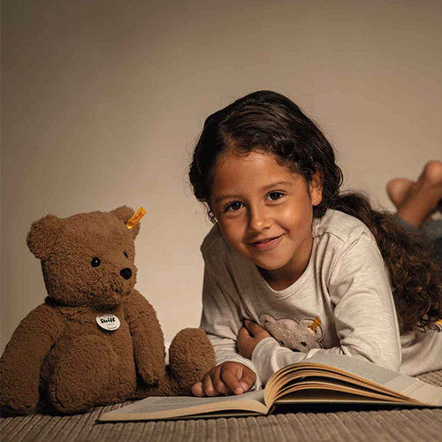 Een meisje lacht naar de camera terwijl ze naast een Steiff pluchen teddybeer en een open boek ligt.