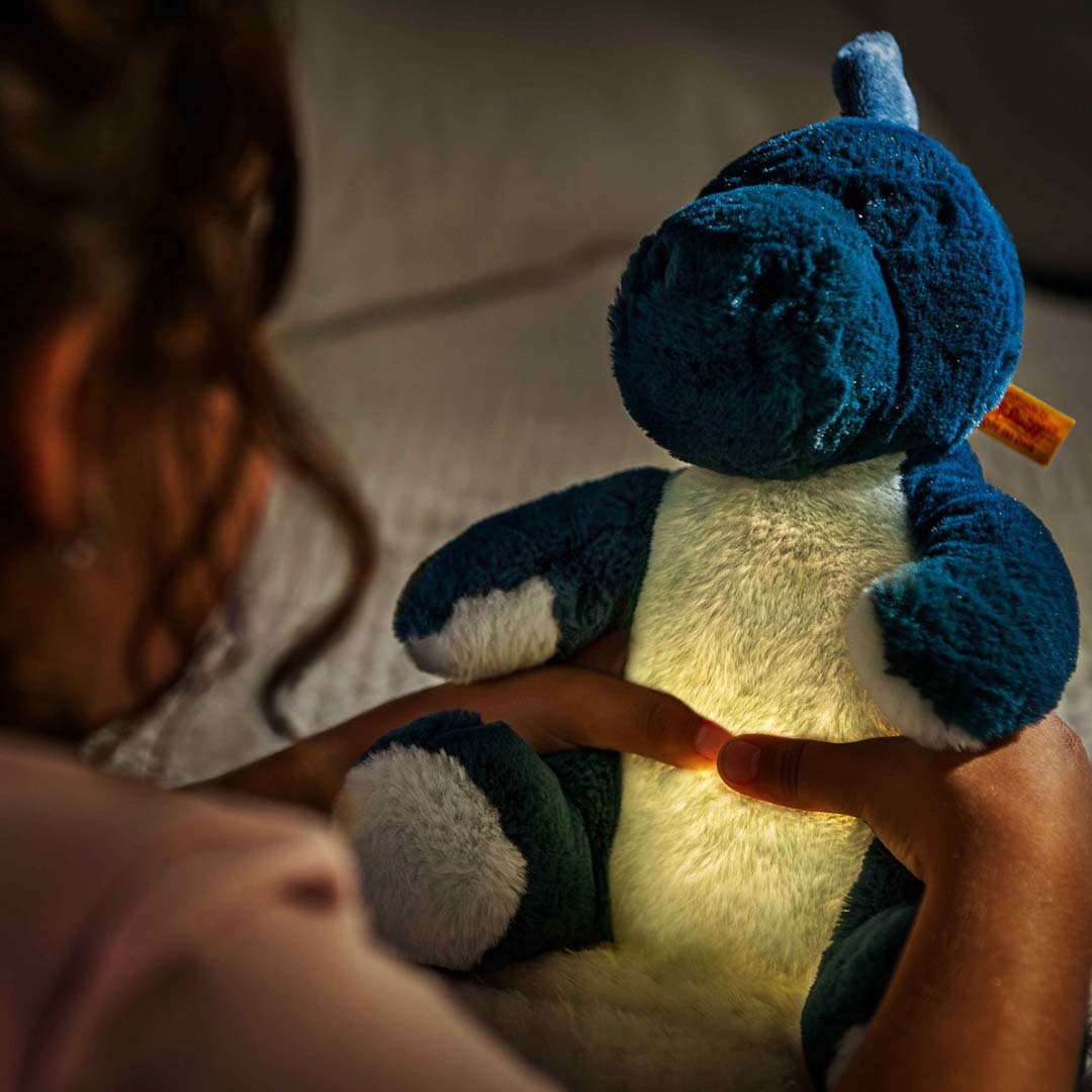 Een kind houdt de Steiff knuffeldinosaurus Spott vast, waarvan de buik oplicht als nachtlampje in het donker.