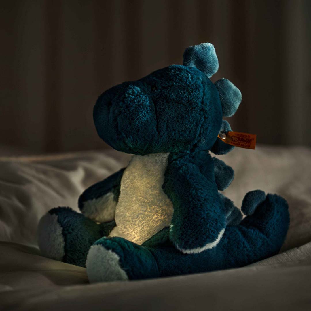 De Steiff knuffeldinosaurus Spott zit op een bed en licht op in het donker, gezien vanaf de achterkant.