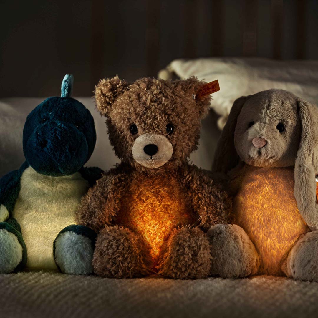 Drie Steiff knuffels, een blauwe dinosaurus, een bruine beer en een grijze konijn, zitten naast elkaar en lichten op in het donker.