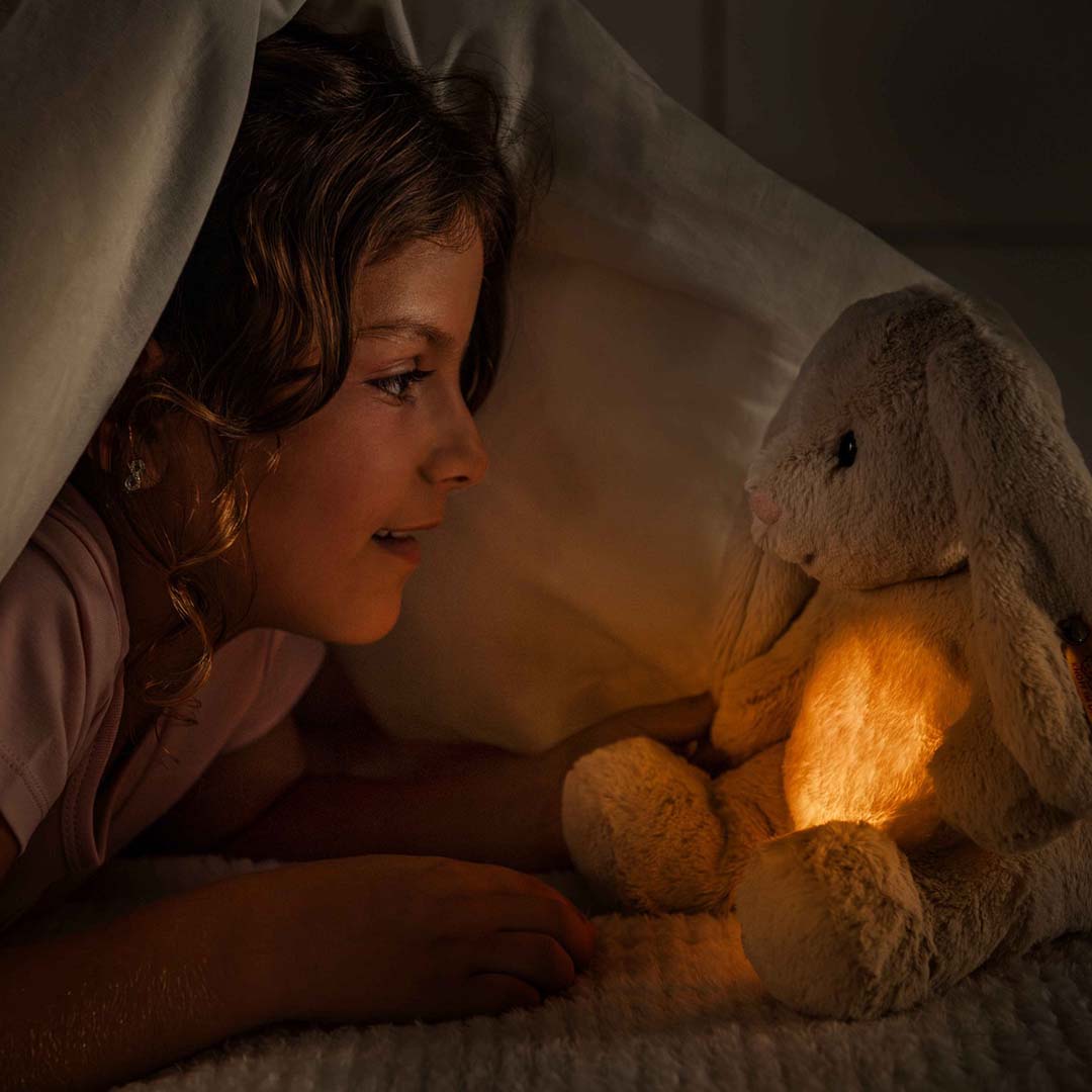 Een meisje kijkt in bed naar het Steiff pluchen knuffelkonijn Hoppi, waarvan de buik oplicht als nachtlampje.