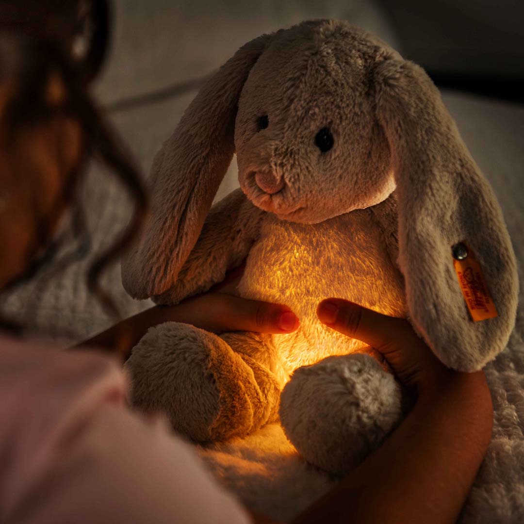 Kinderhanden houden het Steiff pluchen knuffelkonijn Hoppi vast, waarvan de buik warm oranje oplicht als nachtlampje.