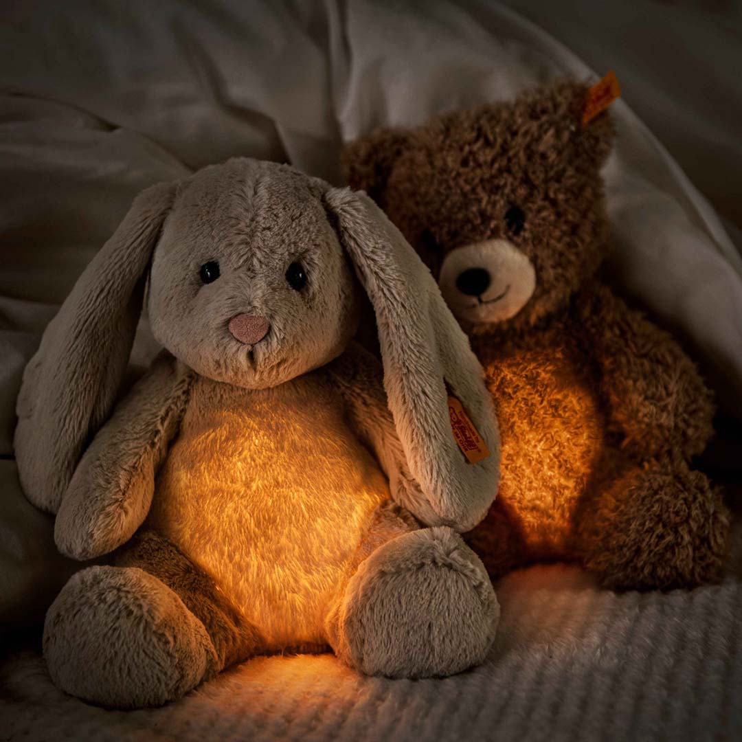 Een oplichtend pluchen konijn en een oplichtende Steiff teddybeer zitten samen op een bed in het donker.