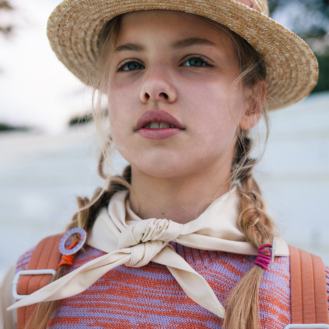Portret van een kind met een strohoed en een oranje Sticky Lemon haarelastiek met gestreepte ring in een vlecht.
