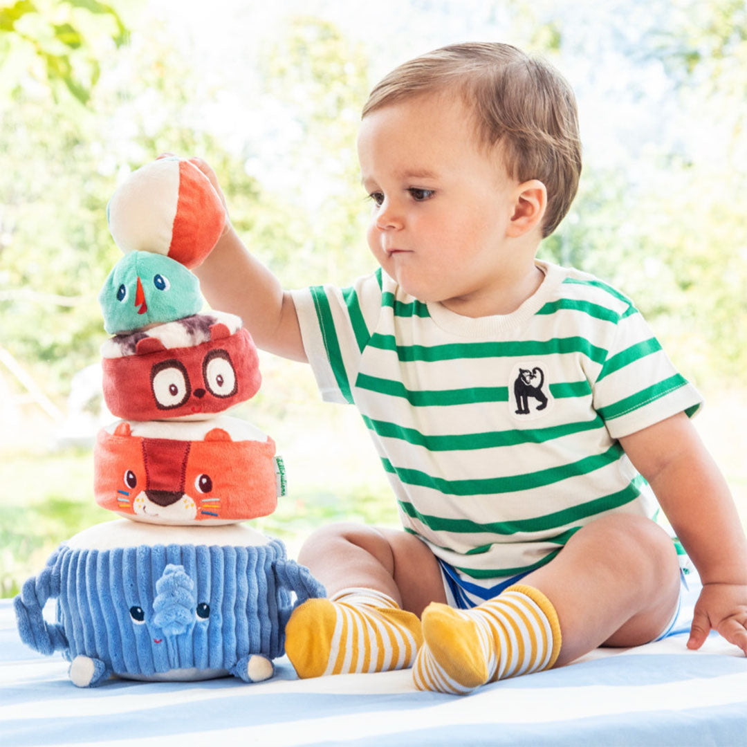 Een baby speelt met de Lilliputiens stapeltoren van zachte dierenblokken, zittend op een blauw-wit gestreepte ondergrond.