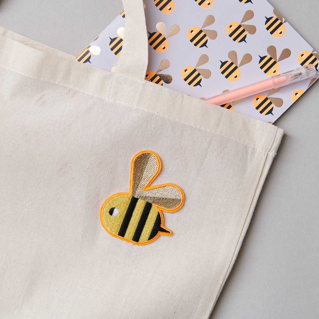 Een opstrijkpatch van een bij op een lichte stoffen tas, met een notitieboekje en pen ernaast.