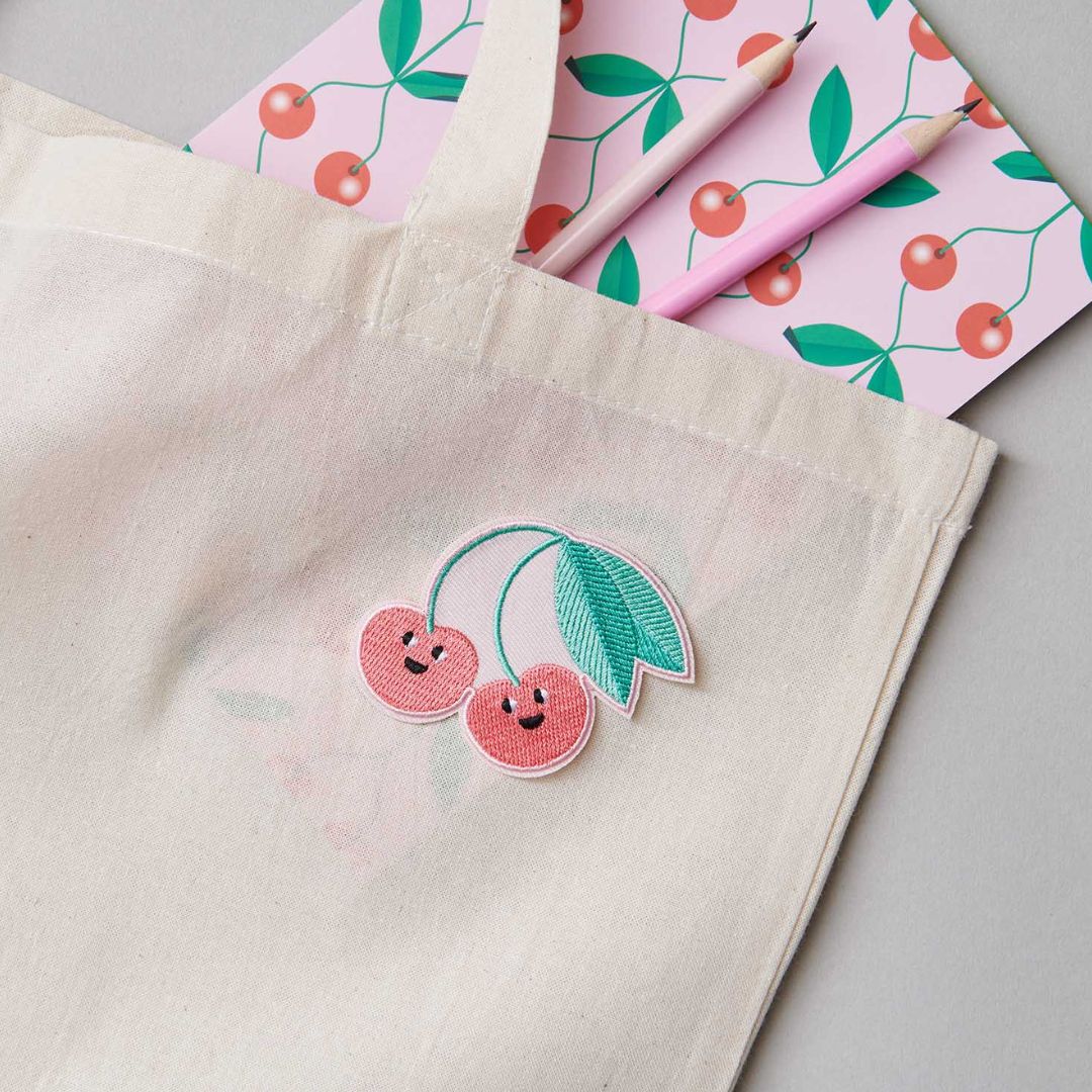 De vrolijke kersen strijkpatch is bevestigd op een beige stoffen tas, met een roze notitieboekje en potloden op de achtergrond.
