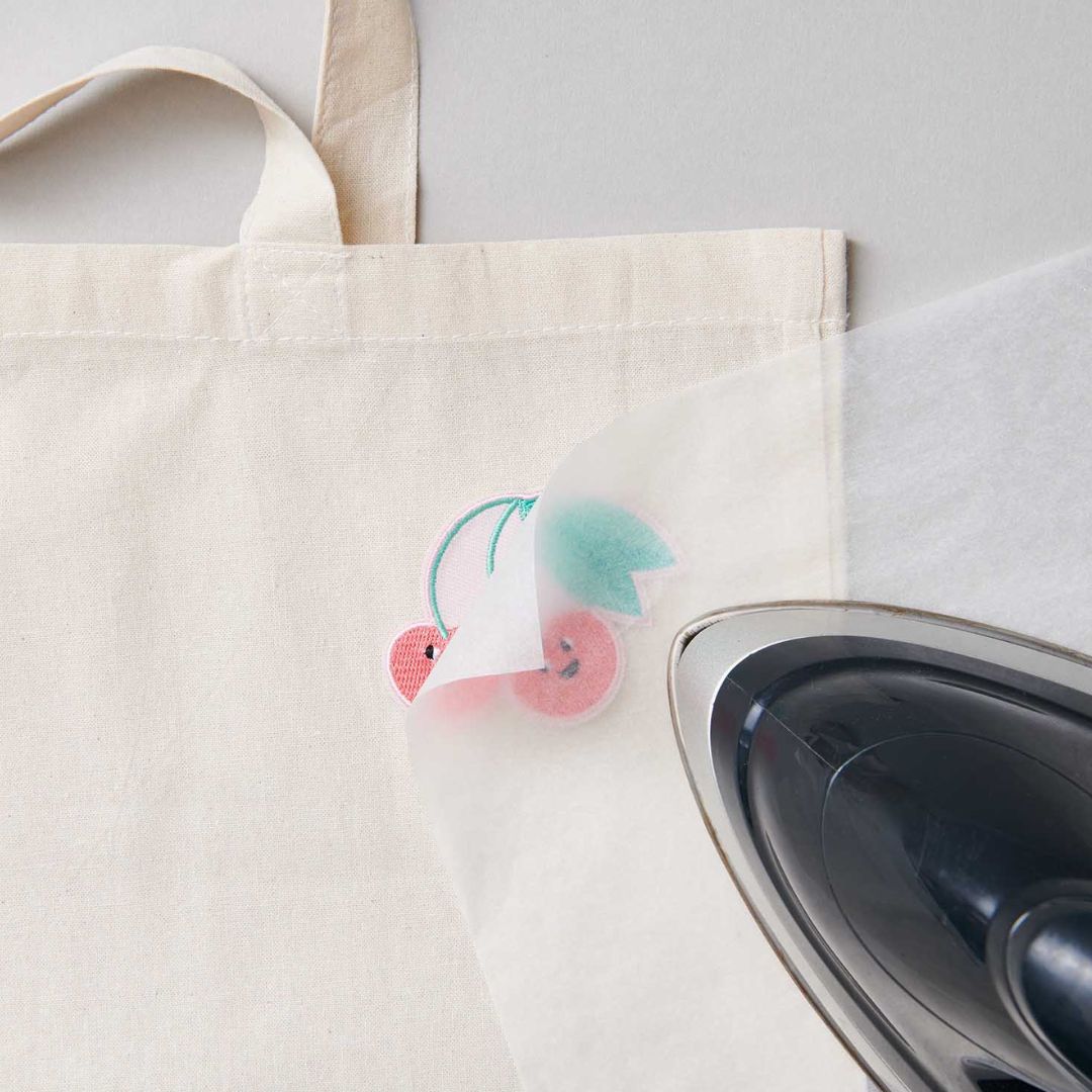 Een strijkijzer bevestigt de kersen strijkpatch met een beschermvel op een beige stoffen tas.