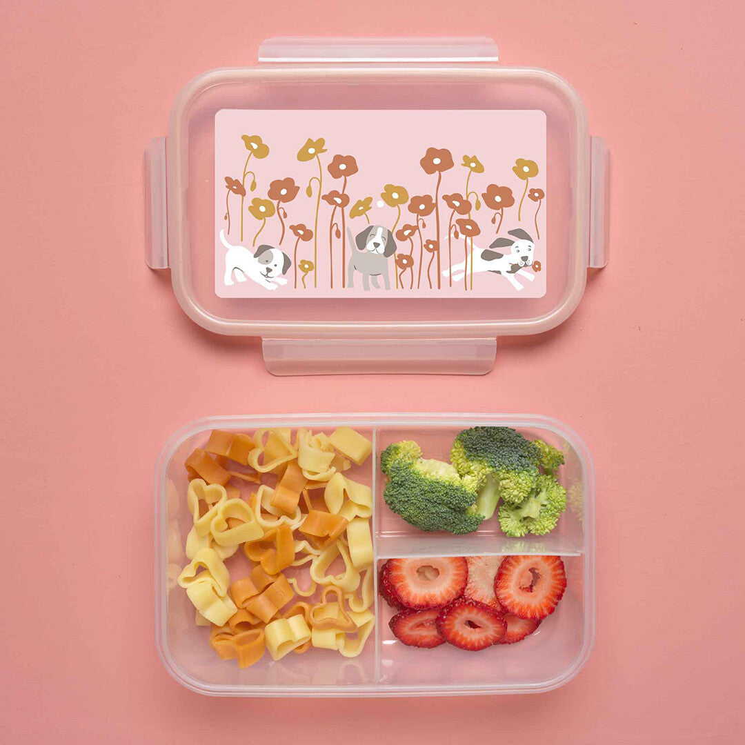 Open bento lunchbox met drie vakken gevuld met pasta, broccoli en aardbeien, en de deksel met puppies en bloemen ernaast.