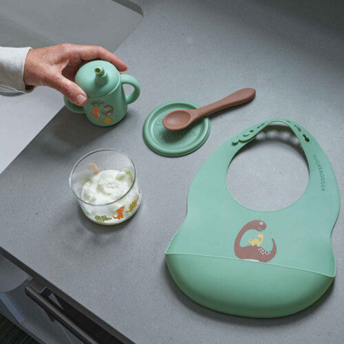 Een sugarbooger babyvoedingsset met een groene slab, drinkbeker, lepel en schaaltje met dinosauruspatroon op een aanrecht.
