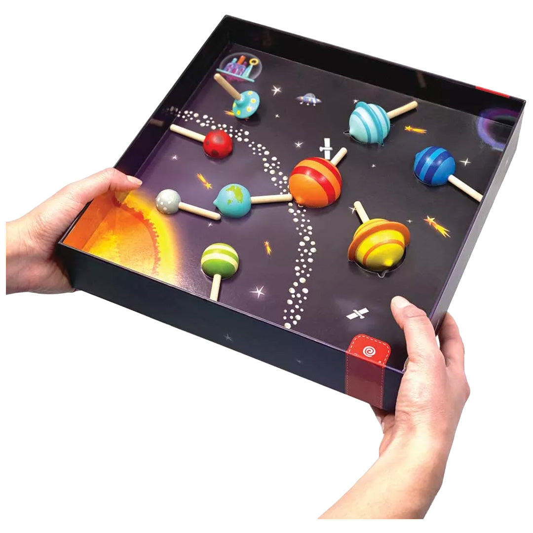 Svoora behendigheidsspel Spinnende Planeten, met twee handen vastgehouden, toont kleurrijke houten tolletjes op een donker speelbord met ruimte-thema.