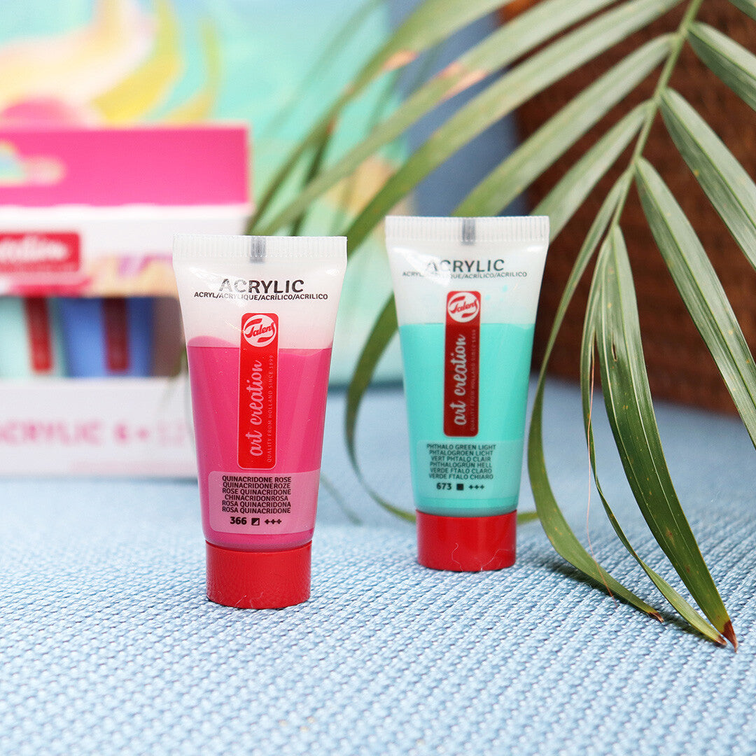 Twee tubes Talens Art Creation acrylverf in roze en turquoise staan op een blauwe ondergrond met een palmblad op de achtergrond.