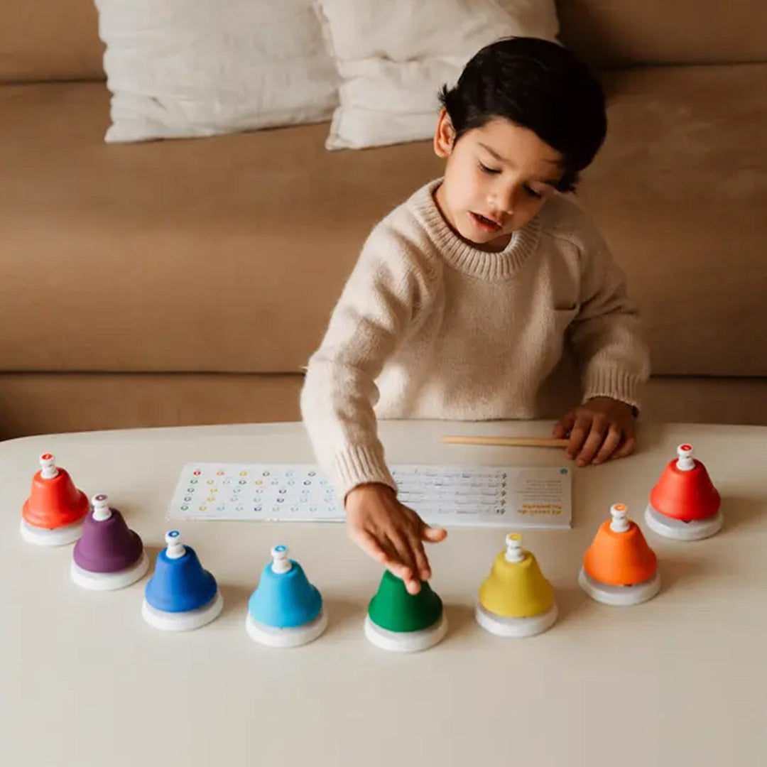 Een kind zit aan tafel en speelt met de 8 kleurrijke muziekklokken, met een muziekboekje voor zich.