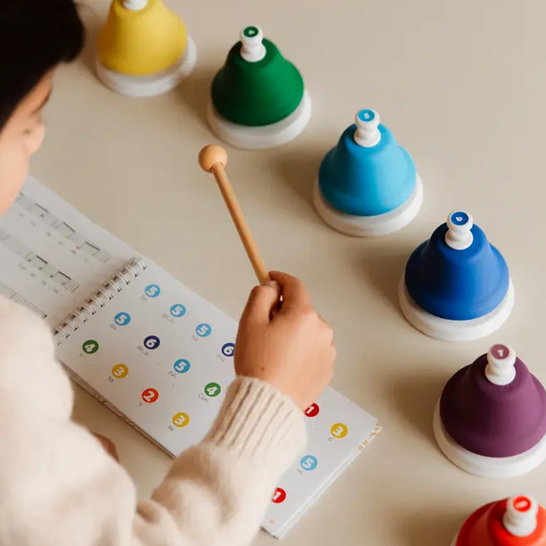 Een kind houdt een houten mallet vast om een van de kleurrijke muziekklokken te bespelen, met een open muziekboek ernaast.