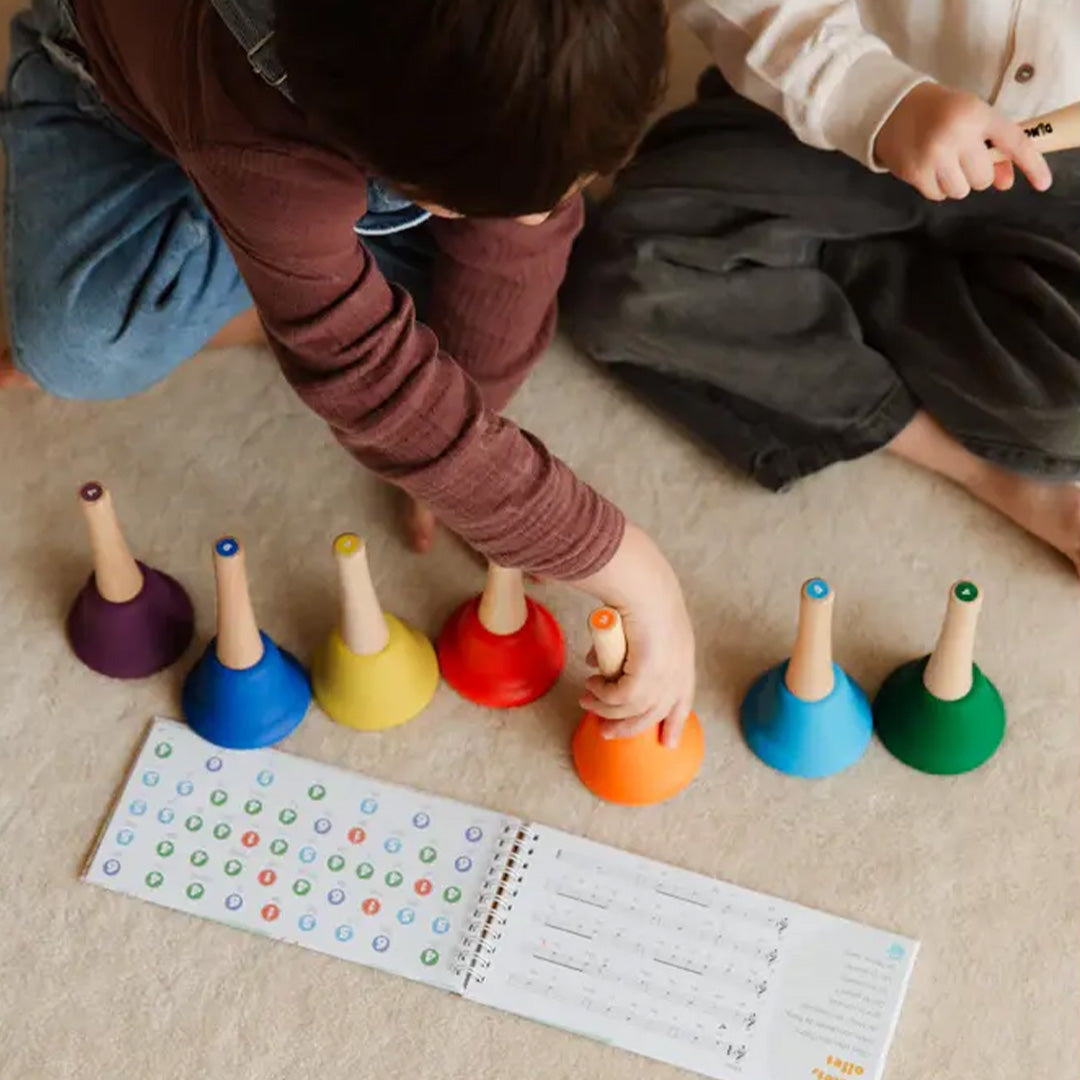 Kinderhanden spelen met de tambu Ding Dang Bú regenboog handbellen en een muziekboekje op de vloer.