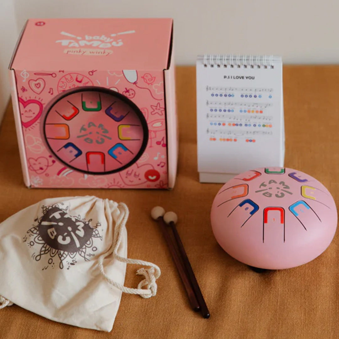 Een set met een roze tambu tongtrommel, de verpakking, een muziekboekje, een opbergzak en twee drumstokken.
