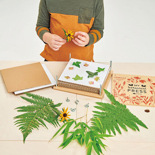 Een kind legt een gele bloem in de open Tender Leaf Toys bloemenpers, omringd door bladeren en bloemen.