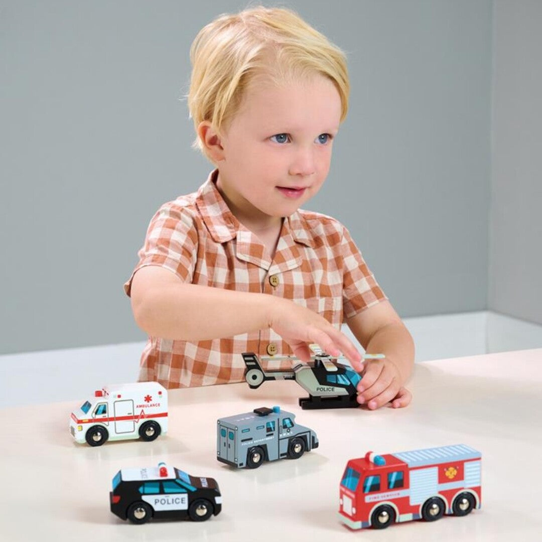 Een kind speelt met de Tender Leaf Toys houten hulpverleningsvoertuigen, waaronder een helikopter en brandweerwagen, op een tafel.