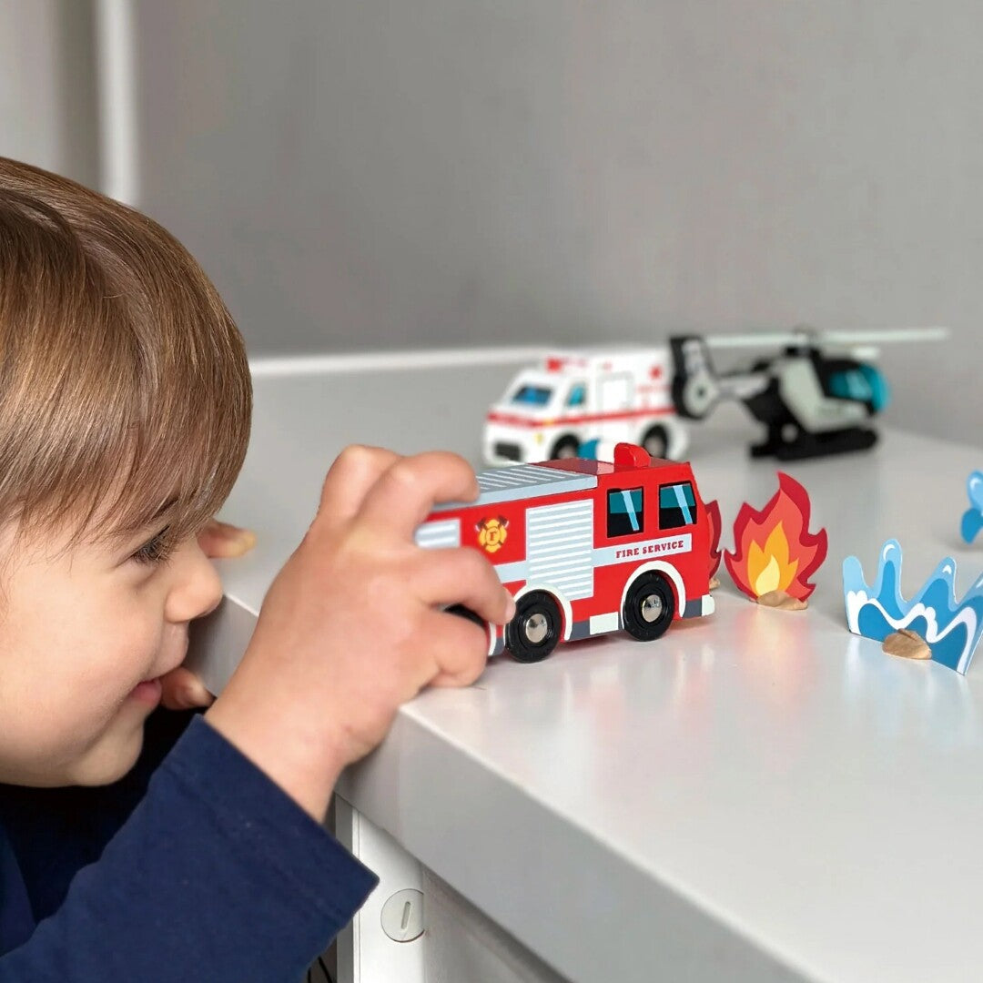 Een kind speelt met een Tender Leaf Toys houten brandweerwagen en houten vlammen en water speelstukken op een tafel.