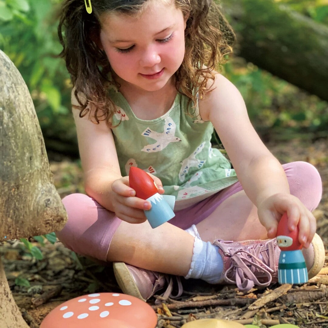 Een meisje speelt buiten met twee houten kabouterfiguren en paddenstoelen.