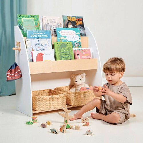 Een peuter speelt met houten speelgoed voor de Tender Leaf Toys witte houten kinderboekenkast gevuld met boeken en manden.