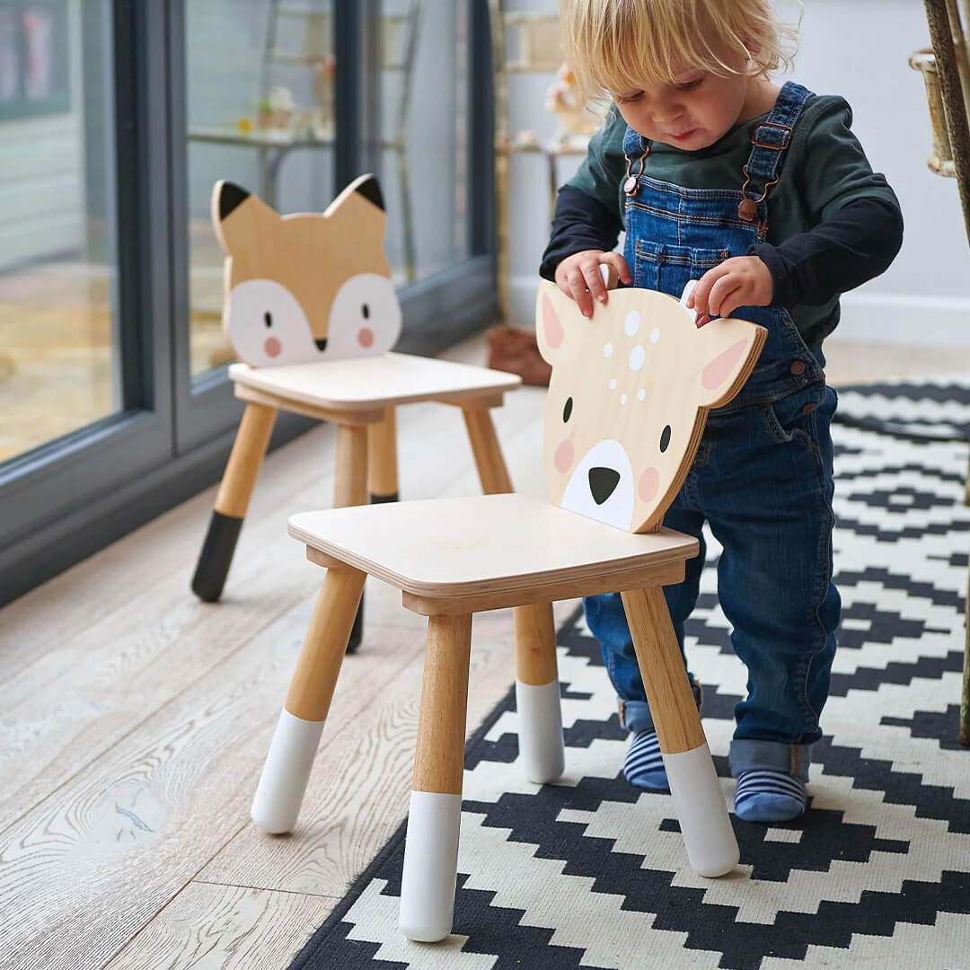 Een peuter staat bij een Tender Leaf Toys houten kinderstoel met hert-rugleuning, met een vossenstoel op de achtergrond.