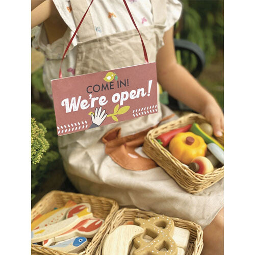 Kind met een 'Open' bordje en drie mandjes Tender Leaf Toys houten speelgoedvoedsel, waaronder brood, vis en groenten.