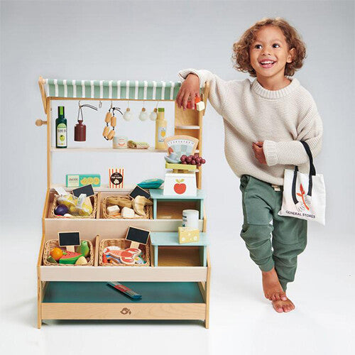 Een lachend kind staat naast de Tender Leaf Toys houten speelgoed marktkraam gevuld met speelgoedvoedsel.