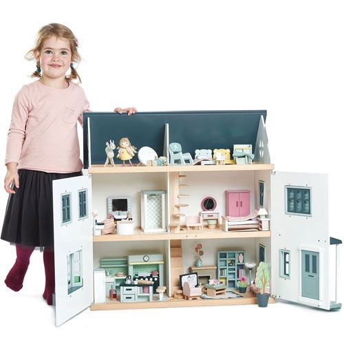 Een lachend meisje staat naast een groot, gemeubileerd Tender Leaf Toys poppenhuis.