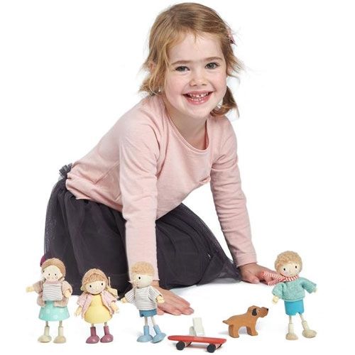 Een lachend meisje knielt naast een set Tender Leaf Toys poppenhuispoppen, een houten hond en een skateboard.