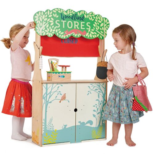 Twee meisjes spelen met het Tender Leaf Toys houten winkeltje, dat gesloten is met een rood gordijn.