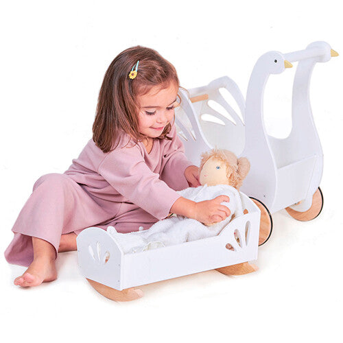 Een meisje speelt met een pop in een witte houten poppenwieg, naast een witte houten poppenwagen met zwanenthema.