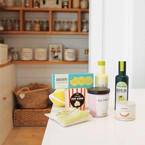 Een selectie Tender Leaf Toys houten speelgoedboodschappen, waaronder crackers, popcorn en olijfolie, op een witte toonbank in een speelkeuken.