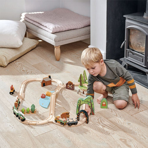 Een jongen speelt met de tender leaf toys houten treinbaan dennenbos, zittend naast een houtkachel.