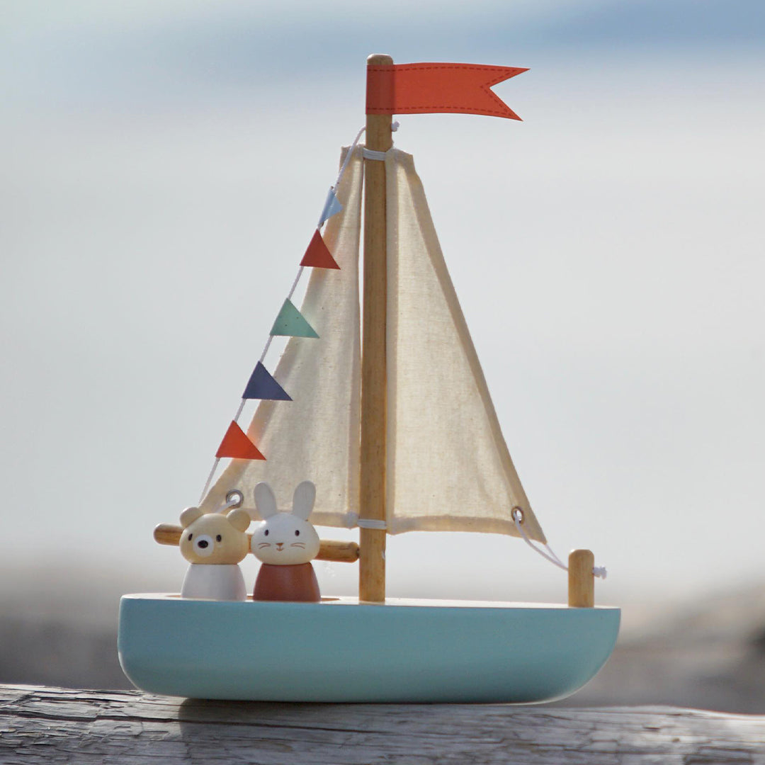 Tender Leaf Toys houten speelgoedzeilboot met een beer en konijn figuur aan boord, staand op een houten balk met een vage waterachtergrond.