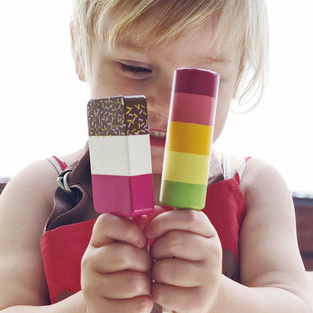 Een kind houdt twee kleurrijke houten speelgoedijsjes met sprinkles en strepen vast.