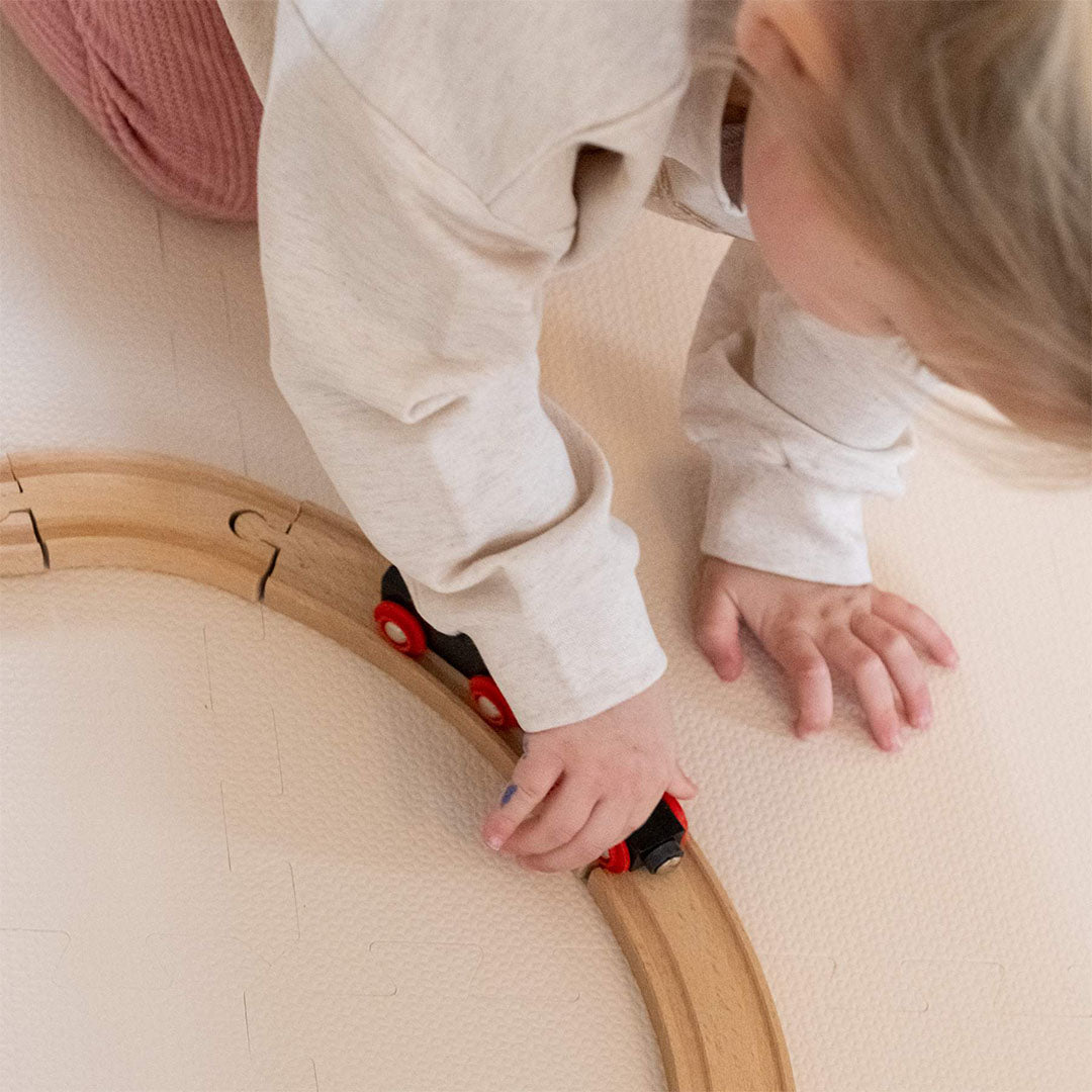Detail van kinderhanden die een houten trein op een spoorbaan bewegen, liggend op de beige foam speelmat.