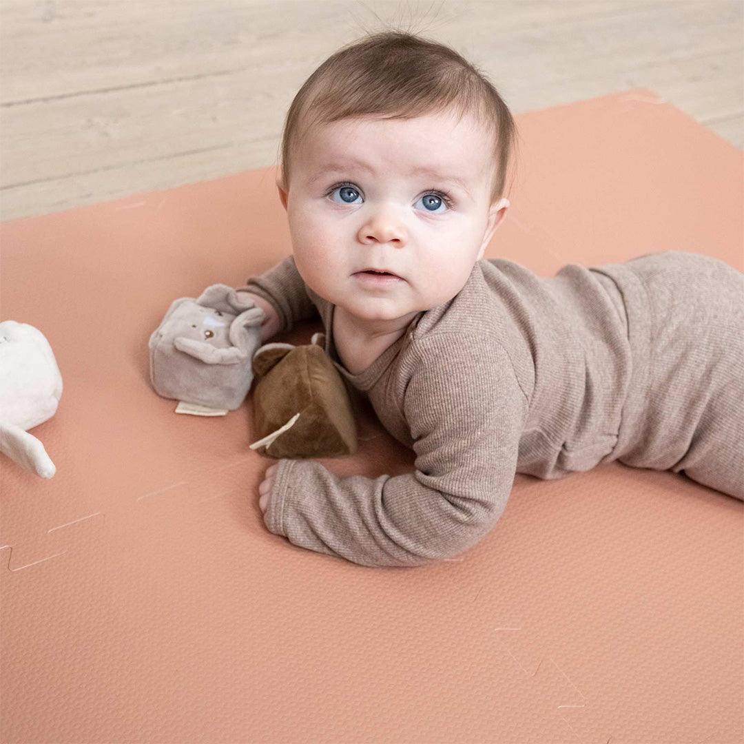 Een baby ligt op de buik op de rood-bruine that's mine foam speelmat met speelgoed.