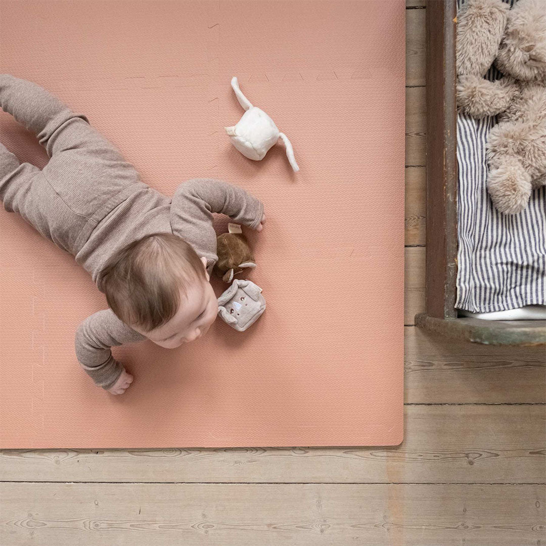 Een baby ligt op de rug op de rood-bruine that's mine foam speelmat en speelt met een knuffeltje.