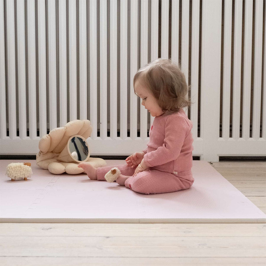 Een baby zit op een roze foam speelmat en kijkt naar een schelpvormig speeltje met spiegel.