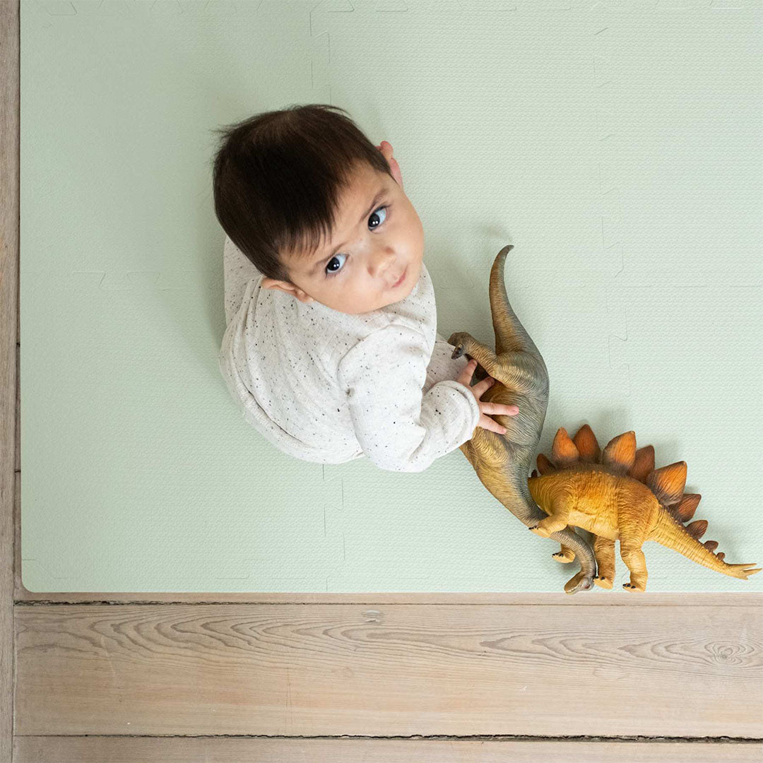 Een baby ligt op de that's mine sage green foam speelmat, met twee dinosaurus speelgoedfiguren ernaast.