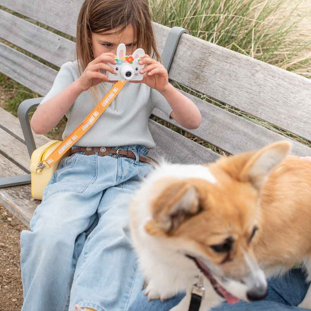 Een kind zit op een bankje en fotografeert met de The Zoofamily kindercamera in de vorm van een konijn, met een hond naast zich.