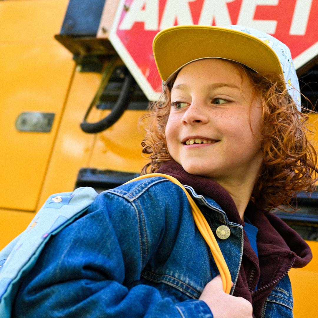 Een lachend kind met rood haar draagt de the cotton cloud gym- en zwemtas en een bijpassende pet, met een schoolbus op de achtergrond.