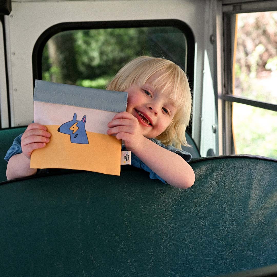 Een lachend kind houdt het the cotton cloud herbruikbaar lunchzakje met funky honden vast, zittend in een bus.