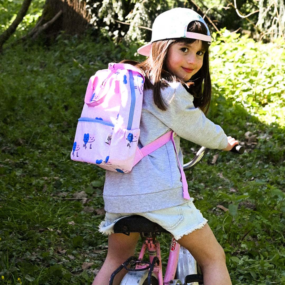 Een meisje draagt de roze kleuterrugzak met dancing hearts patroon op een fiets in het bos.