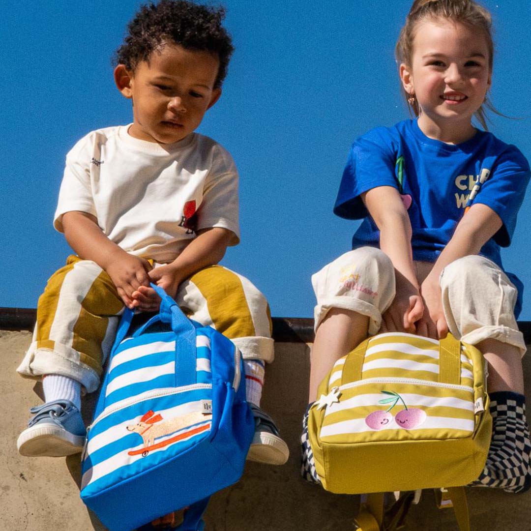 Twee kinderen zitten met The Cotton Cloud kleuterrugzakken, één blauw-wit gestreept met een hond en één geel-wit gestreept met kersen.