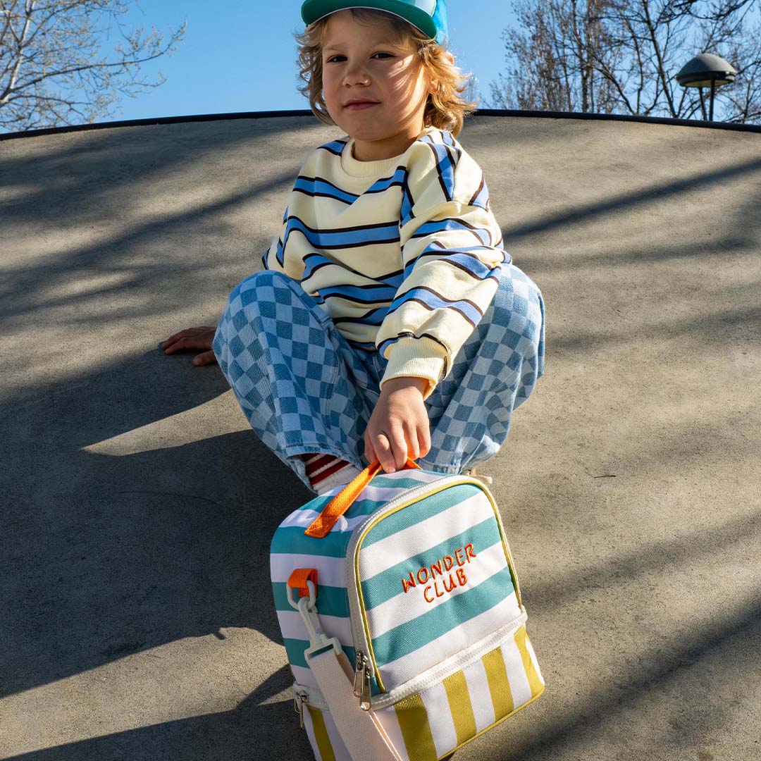 Een kind zit met de the cotton cloud gestreepte koeltas met 'Wonder Club' tekst voor zich op de grond.
