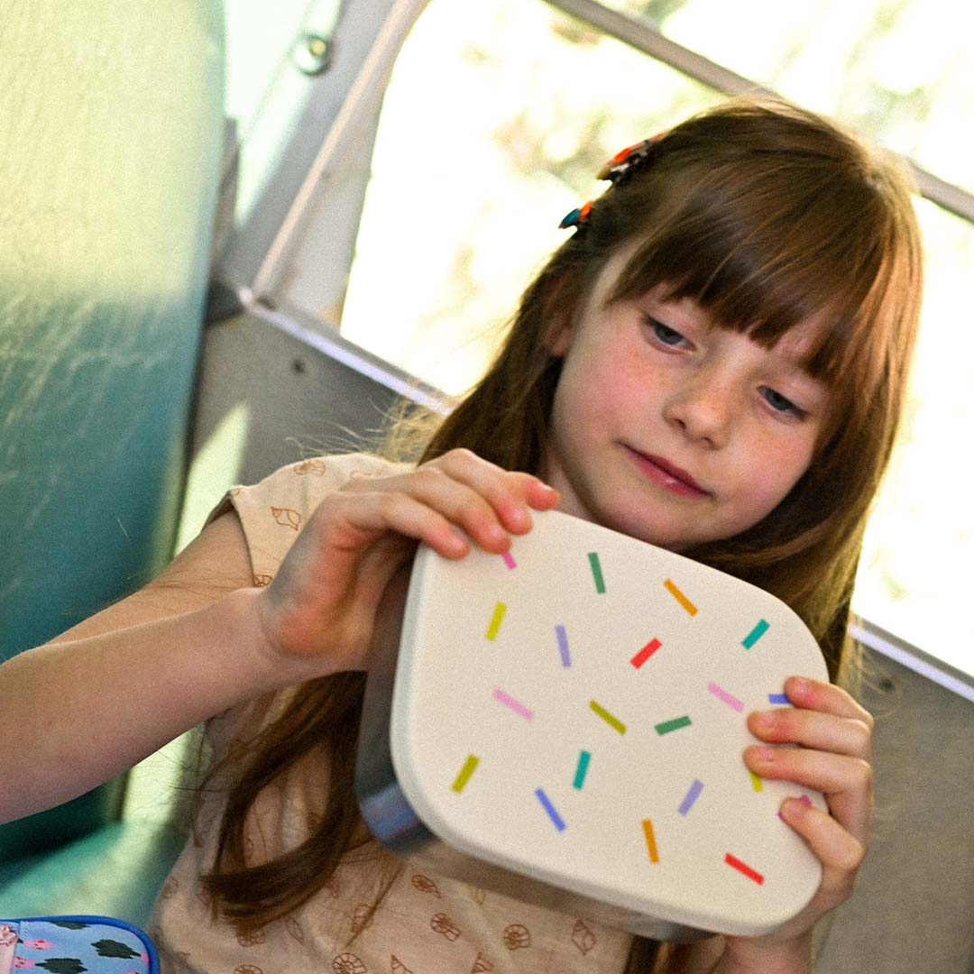 Een kind houdt een gesloten kinderbroodtrommel met confetti deksel vast.